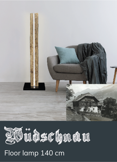 A wooden floor lamp with an illuminated strip stands next to a grey armchair with cushion and blanket in a modern living room. Text: &ldquo;W&uuml;dschnau floor lamp 140 cm.&rdquo; Black-and-white photo of a chalet.